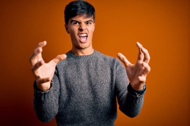 İzole edilmiş turuncu arka plan üzerinde duran genç ve yakışıklı bir adam öfkeyle haykırıyor, elleri boğmaya çalışıyor, deli gibi bağırıyor.