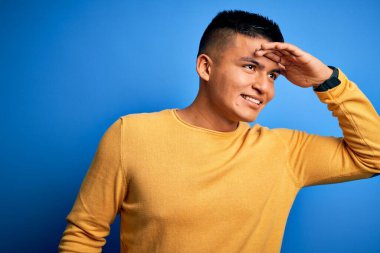 Soyutlanmış mavi arka plan üzerinde sarı günlük süveter giyen genç, yakışıklı latin adam çok mutlu ve tebessüm ediyor. Konsept aranıyor.