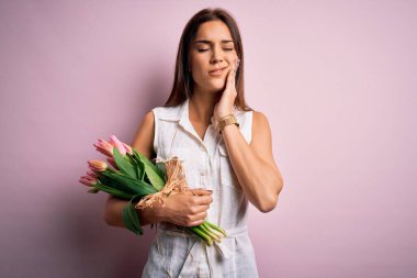 Genç, güzel, esmer bir kadın pembe arka planda bir buket lale çiçeği tutuyor diş ağrısı ya da diş hastalığından dolayı acı dolu bir ifadeyle dudaklarına dokunuyor. Dişçi konsepti.