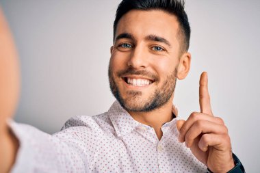 Gömlek giyen genç yakışıklı bir adam kameranın önünde özçekim yapıyor beyaz arka planda bir fikir ya da soru işareti ile şaşırıyor mutlu bir yüzle, bir numara