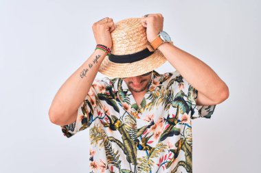 Havai gömleği ve yaz şapkası takan genç yakışıklı bir adam izole bir arka planda acı ve migren yüzünden çaresiz ve stresli bir baş ağrısından muzdarip. Ellerini başının üstüne koy..