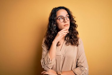 Kıvırcık saçlı, çizgili tişörtlü, sarı arka planda gözlüklü, eli çenesinde soru soran, düşünceli bir kadın. Düşünceli bir yüzle gülümsüyor. Şüphe kavramı.