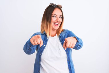 İzole edilmiş beyaz arka planda duran kot gömlek giyen güzel bir kadın seni ve kamerayı parmaklarıyla gösteriyor, mutlu ve mutlu gülümsüyor.