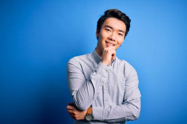 Genç yakışıklı Çinli bir adam mavi arka plan üzerinde dikilip kolları çapraz ve elleri çene üzerinde gülümseyen bir şekilde kameraya bakıyor. Olumlu düşünüyorum..