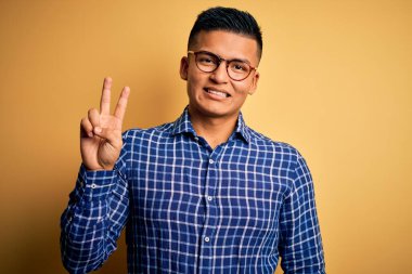 Sarı arka planda gözlüklü, sıradan tişörtlü genç, yakışıklı Latin adam kameraya bakıp zafer işareti yapıyor. İki numara..