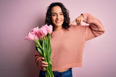 Genç, kıvırcık saçlı, pembe lale buketi tutan, kendine güvenen, gülümseyen, kendini gururlu ve mutlu parmaklarıyla gösteren romantik bir kadın..