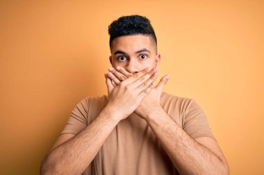 Izole sarı arka plan üzerinde duran rahat t-shirt giyen genç yakışıklı adam hata için elleri ile ağız kapsayan şok. Gizli kavram.