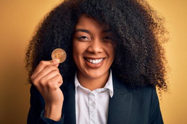 Afro saçlı, sarı arka planda bitcoin tutan, gülümseyen, kendine güvenen gülümseyen, dişlerini gösteren Afro Amerikalı genç bir iş kadını.