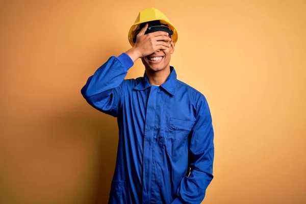 青い制服とセキュリティヘルメットを身に着けている若いハンサムなアフリカ系アメリカ人労働者の男性は笑顔と驚きのために目をカバー顔に手で笑っています ブラインドコンセプト — ストック写真