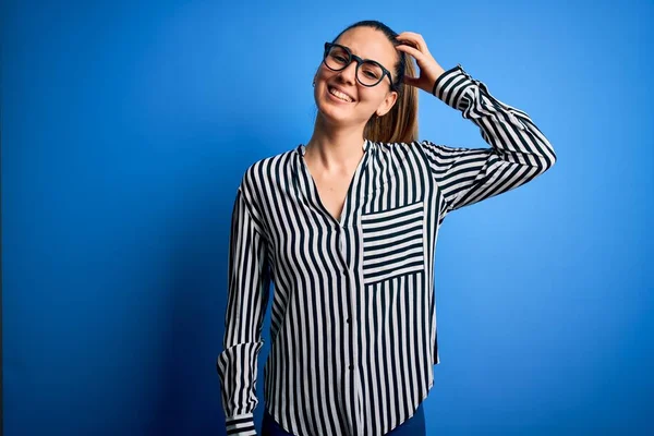 Beautiful blonde woman with blue eyes wearing striped shirt and glasses over blue background confuse and wonder about question. Uncertain with doubt, thinking with hand on head. Pensive concept.