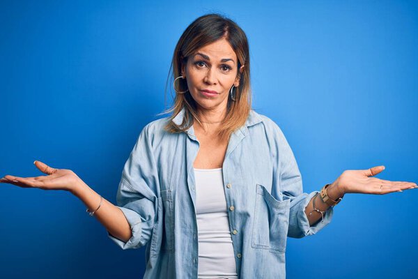 Middle age beautiful woman wearing casual shirt standing over isolated blue background clueless and confused with open arms, no idea concept.