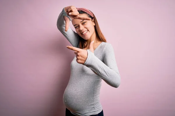 孤立したピンクの背景に赤ちゃんを期待して若い美しいティーンエイジャーの女の子は幸せな顔を持つ手と指でフレームを作り笑顔 創造性と写真の概念 — ストック写真