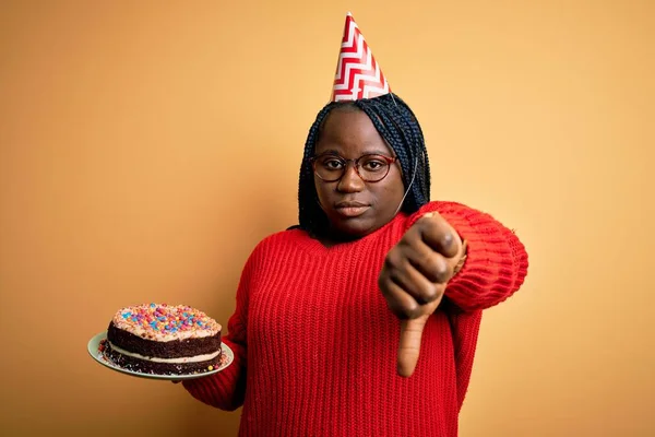 非洲裔美国人 身材的女人头戴一顶生日帽 满脸怒容 头戴黄色背景的蛋糕 脸上挂着厌恶大拇指的负面标志 被拒绝的念头 — 图库照片