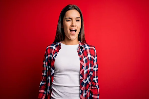 Young beautiful brunette woman wearing casual shirt standing over isolated red background winking looking at the camera with sexy expression, cheerful and happy face.