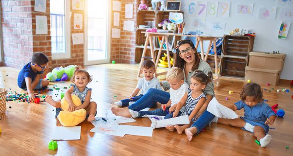 Beautiful teacher and group of toddlers sitting on the floor drawing using paper and pencil around lots of toys at kindergarten