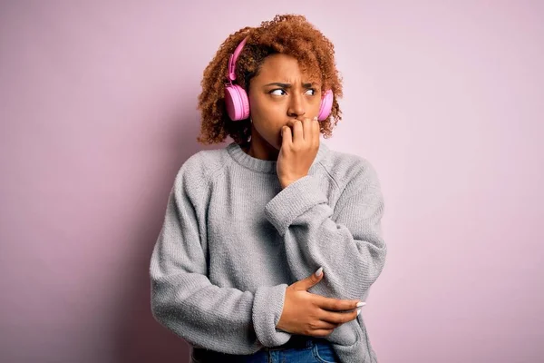Young African American afro woman with curly hair listening to music using pink headphones looking stressed and nervous with hands on mouth biting nails. Anxiety problem.
