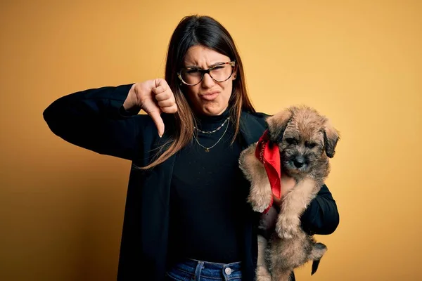 Young beautiful brunette woman holding cute puppy pet over isolated yellow background with angry face, negative sign showing dislike with thumbs down, rejection concept