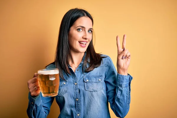 年轻的女人 蓝眼睛 喝着一罐啤酒 站在孤立的黄色背景上 微笑着看着摄像机 手指头在做胜利的标志 第二点 — 图库照片