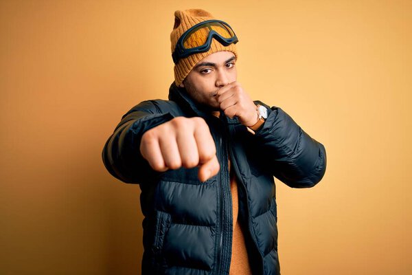 Молодой бразильский лыжник в снежной спортивной одежде и лыжных очках на жёлтом фоне бьет кулаком в драку, агрессивное и злое нападение, угроза и насилие
