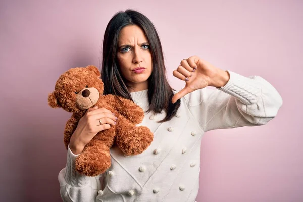 Young brunette woman with blue eyes hugging teddy bear stuffed animal over pink background with angry face, negative sign showing dislike with thumbs down, rejection concept