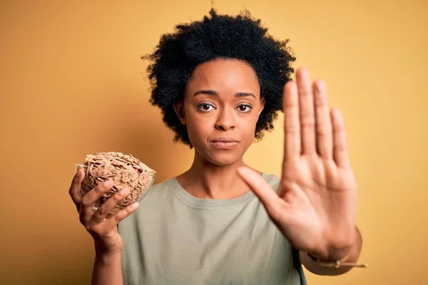 Young African American afro woman with curly hair holding bowl with cornflakes cereals with open hand doing stop sign with serious and confident expression, defense gesture