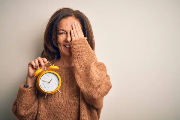 中年ブルネット女性保持古典的な目覚まし時計上の隔離された背景カバー1目に手で 自信を持って笑顔上の顔と驚きの感情 — ストック写真