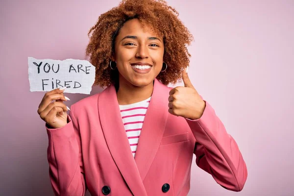 Young African American afro woman with curly hair holding papaer with you are fired message happy with big smile doing ok sign, thumb up with fingers, excellent sign