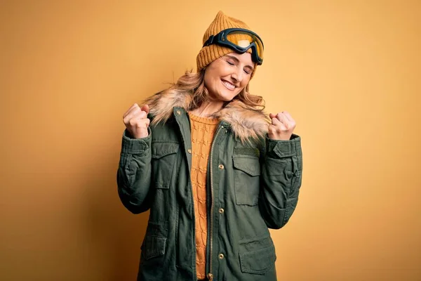 中年漂亮的金发滑雪者女人穿着雪地运动鞋和滑雪护目镜 非常高兴而兴奋地举起双臂做获奖者的手势 微笑着 尖叫着要成功 庆祝概念 — 图库照片