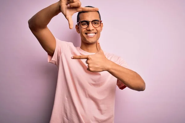 ピンクの背景にカジュアルなTシャツと眼鏡を身に着けているハンサムなアフリカ系アメリカ人男性は 幸せな顔をした手と指でフレームを作り笑顔 創造性と写真の概念 — ストック写真