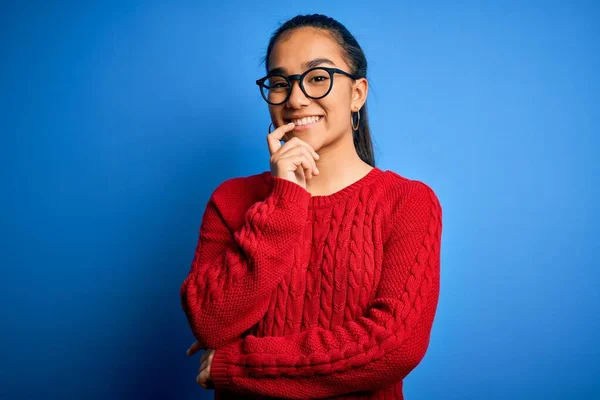 Young beautiful asian woman wearing casual sweater and glasses over blue background looking confident at the camera with smile with crossed arms and hand raised on chin. Thinking positive.