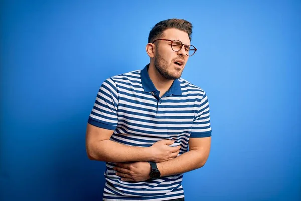 Young man with blue eyes wearing glasses and casual striped t-shirt over blue background with hand on stomach because nausea, painful disease feeling unwell. Ache concept.