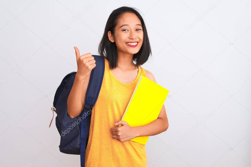 Young chinese student woman wearing backpack holding book over isolated white background happy with big smile doing ok sign, thumb up with fingers, excellent sign