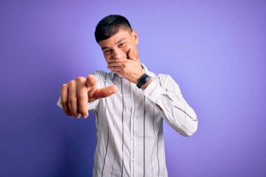 Zarif iş gömleği giyen genç yakışıklı İspanyol adam mor arka planda duruyor sana gülüyor, kamerayı işaret ediyor, ağzının üzerinde utanç ifadesi var.