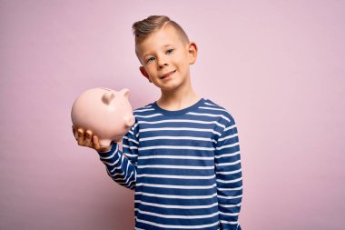 Küçük beyaz çocuk domuz kumbarasına para yatırıyor pembe arka planda servet biriktirirken mutlu bir yüzü olan ve kendine güvenen bir gülümsemeyle dişlerini gösteren