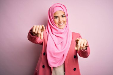 İslami tesettürlü genç ve güzel bir kız pembe arka planda dikiliyor. Seni ve kamerayı parmaklarıyla gösteriyor, mutlu ve mutlu gülümsüyor.