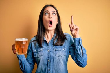 Mavi gözlü genç kadın, izole edilmiş sarı arka planda duran bira kavanozunu içerken şaşkın şaşkın ve şaşkın bir şekilde yukarı bakıyor ve parmaklarıyla işaret ediyor ve kollarını kaldırıyordu..