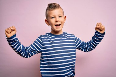 Mavi gözlü, pembe arka plan üzerinde deniz çizgili tişört giyen küçük beyaz bir çocuk kollarını kaldırarak ve açık gözlerle başarıyı kutluyor ve şaşırıyor. Kazanan konsept.