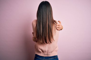 Genç, güzel, esmer bir kadın, pembe arka planda rahat bir süveter giyiyor, mutlu ve pozitif bir şekilde kendine sarılıyor. Sevgi ve özgüven.