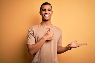 Genç, Afro-Amerikalı, günlük tişörtler giyen, sarı arka planda duran, el hareketi yapan, mutlu ve neşeli gülümseyen yakışıklı bir adam.
