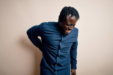 Beyaz arka planda dikilen, sırt ağrısı çeken, eli ile dokunan, kaslı, gündelik gömlekler giyen genç, Afro-Amerikan bir adam.