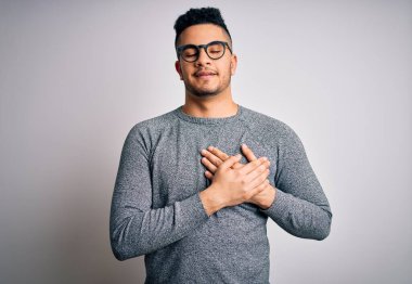 İzole edilmiş beyaz arka planda gözlük ve süveter takan genç yakışıklı adam elleri göğsünde, gözleri kapalı ve yüzünde minnet dolu bir jest ile gülümsüyor. Sağlık kavramı.