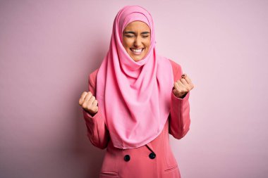 Genç ve güzel esmer iş kadını pembe Müslüman başörtüsü ve iş ceketi giyiyor. Çok mutlu ve heyecanlı. Kazanan jestler yapıyor. Kutlama konsepti.
