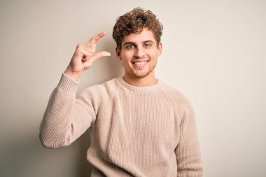 Young blond handsome man with curly hair wearing casual sweater over white background smiling and confident gesturing with hand doing small size sign with fingers looking and the camera. Measure concept.