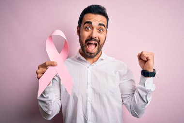 Sakallı, elinde pembe kanser kurdelesi olan genç yakışıklı adam izole edilmiş arka planda gururla bağırıyor, zaferi ve başarıyı kutluyor. Çok heyecanlı, coşkulu bir duygu.