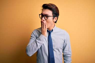 Sarı arka planda gözlük takan ve kravat takan genç yakışıklı Çinli işadamı ağzıyla esnemekten sıkılmış. Huzursuz ve uykulu..