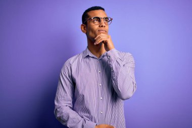 Çizgili gömlek ve mor arka planda gözlük takan yakışıklı Afro-Amerikan erkeği. Elleri çenesinde soru sorarken, düşünceli bir ifade takınıyor. Gülen ve düşünceli bir yüz. Şüphe kavramı.