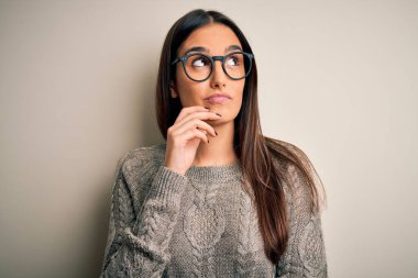 Genç, güzel, esmer bir kadın, beyaz arka plan üzerinde gözlük ve süveter giyiyor, elleri çenesinde soru sormayı düşünüyor, düşünceli bir ifade. Düşünceli bir yüzle gülümsüyor. Şüphe kavramı.