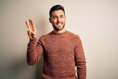 İzole edilmiş beyaz arka plan üzerinde duran sıradan süveter giyen genç ve yakışıklı bir adam üç numaralı parmaklarıyla işaret ederken kendinden emin ve mutlu gülümsüyor..
