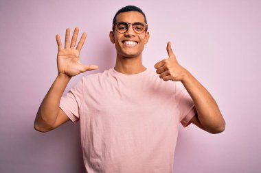 Yakışıklı Afro-Amerikalı adam pembe arka planda günlük tişört ve gözlük takıyor ve altı numarayı gösterirken kendinden emin ve mutlu gülümsüyor..