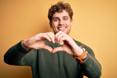 Sarı arka planda yeşil kazak giyen, kıvırcık saçlı, genç, sarışın ve yakışıklı bir adam. Aşkla gülümsüyor. Elleriyle kalp sembolü ve şekli gösteriyor. Romantik konsept.